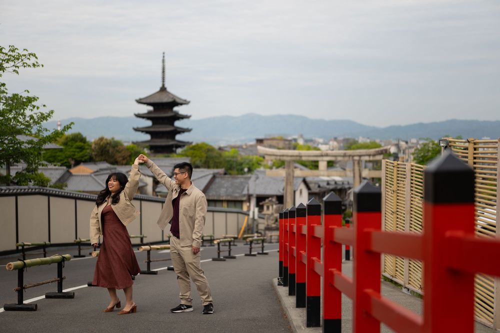 Couple in casual outfits dance on a Kyoto street beside red railings, with a traditional pagoda and mountains in the background.