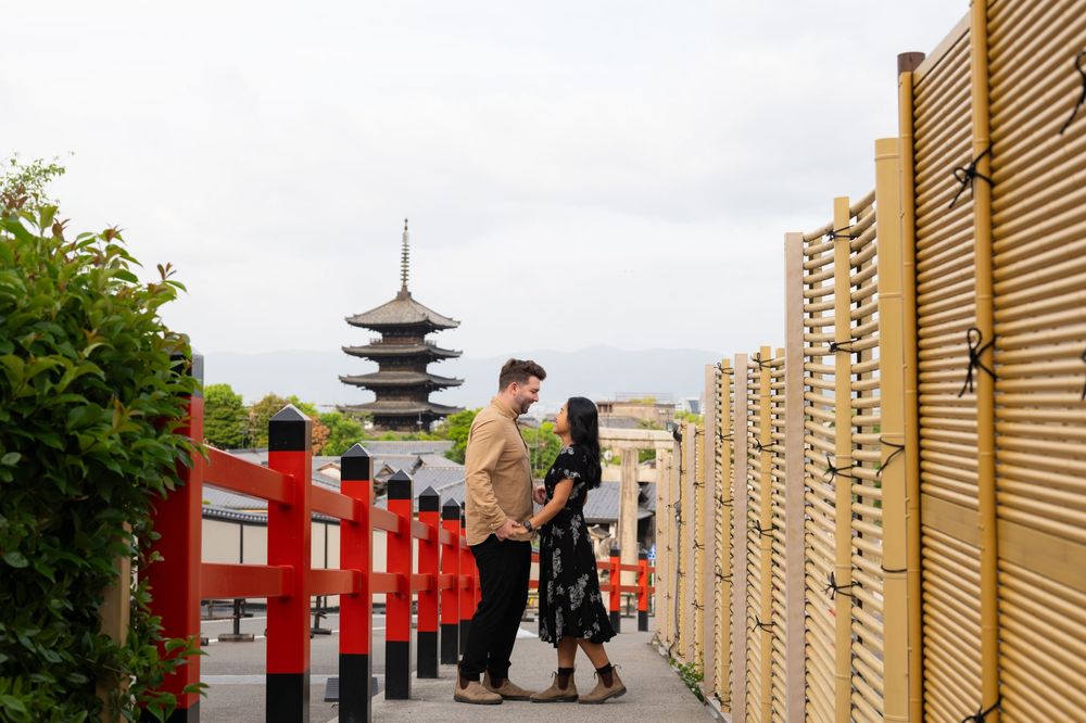 Couple embracing on a stone path beside a red railing and bamboo fence, with a Japanese pagoda rising in the background under a cloudy sky.