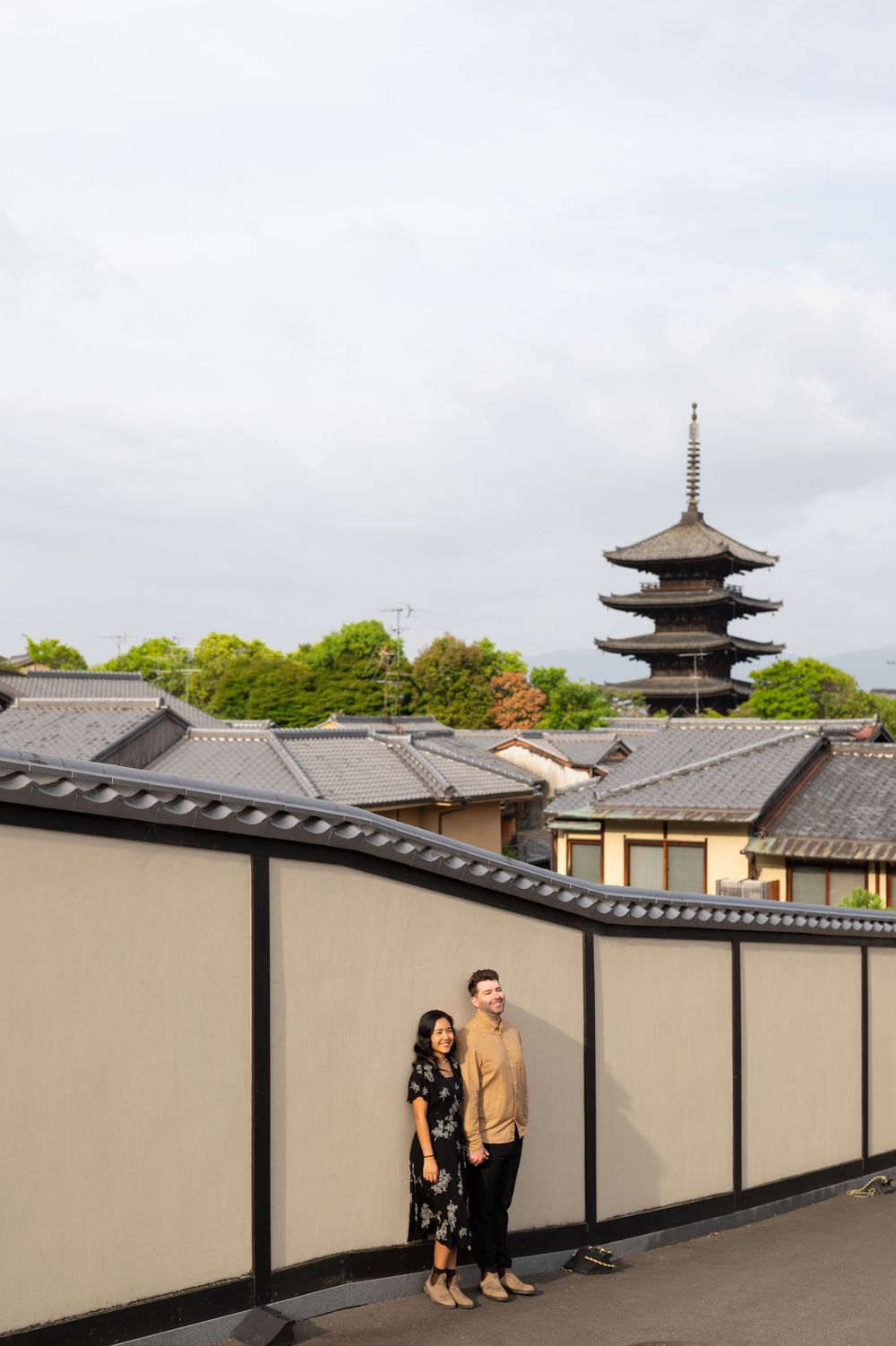 A couple stands on a rooftop terrace beside a cream wall, overlooking traditional tiled roofs and a tall five-story pagoda in Kyoto.