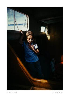 Child in a blue shirt stands in a dimly lit sailboat cabin, touching a window as warm sunlight streams across the wooden interior.