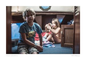 Two children relax inside a cozy sailboat cabin, a boy in a blue graphic t-shirt sitting in front while a girl plays behind him.