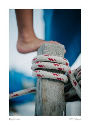 Bare foot resting on a weathered wooden dock post wrapped with white and red rope, with blue sailboat details blurred in the background.