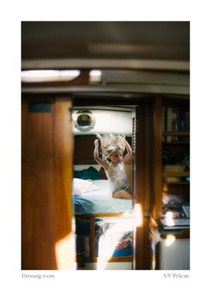 Young blonde child jumping on a bed in a wood-paneled sailboat cabin aboard S/V Pelican, seen through a doorway.