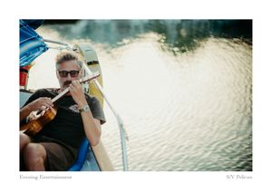 A man in sunglasses plays a ukulele aboard a sailboat in the evening, with sparkling water reflecting light behind him.