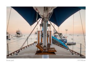View from a sailboat deck at sunset, showing the foredeck, rigging, and blue sail cover with boats anchored in a calm harbor.