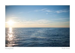 Golden sun low on the horizon casts a shimmering reflection across calm blue Pacific Ocean waters under a soft, cloud-streaked sky.