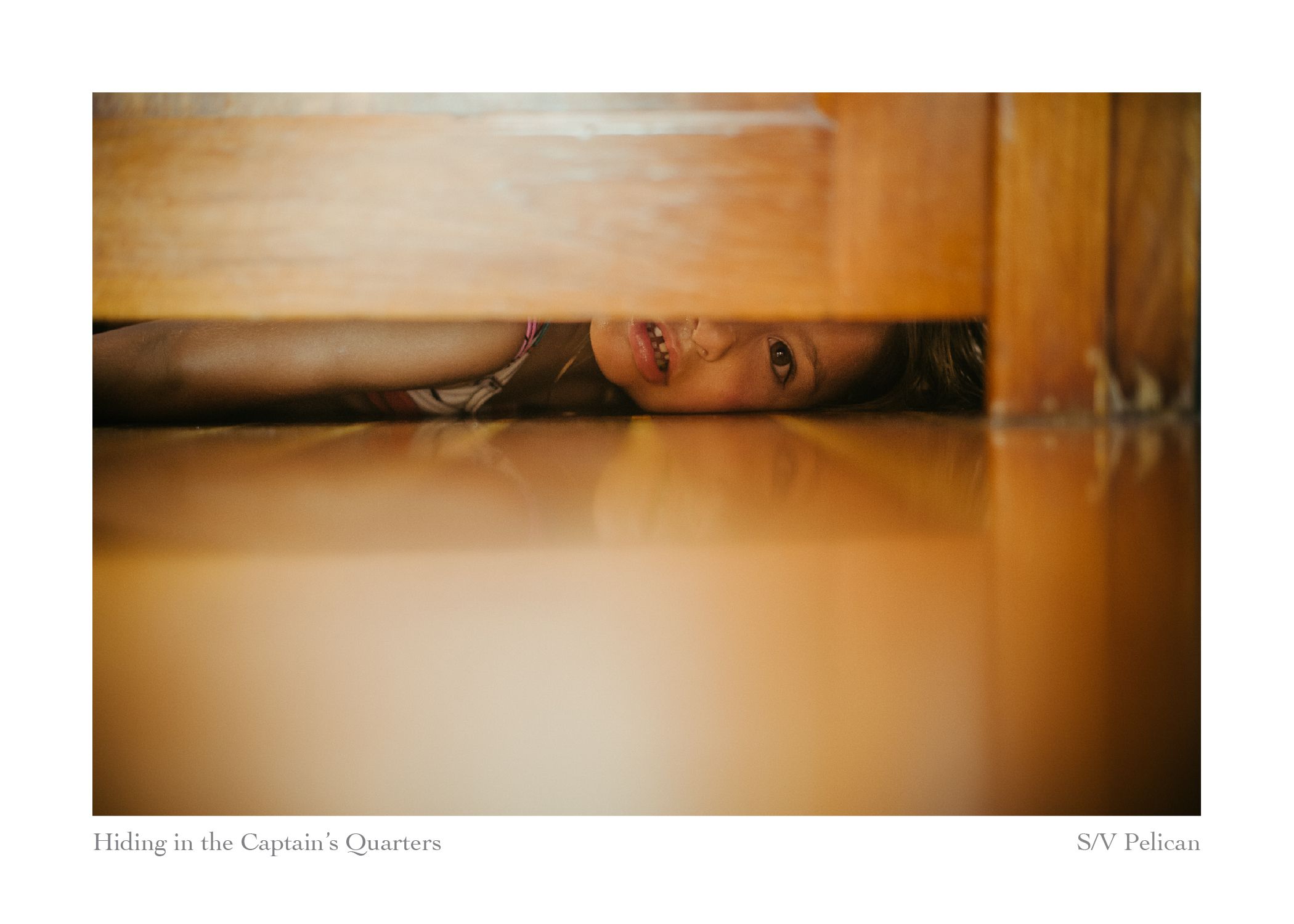 A smiling child peeks out from under a wooden berth, hiding playfully in the captain's quarters of a sailboat.