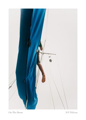 Barefoot sailor standing on a sailboat boom beside a bright blue sail cover, with rigging against a pale sky.