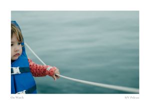Toddler in a blue life jacket and red patterned sleeves holds a white sailboat line while gazing over calm blue-green water.