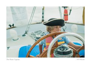 Child in a black pirate hat and life vest steers a sailboat with a wooden helm wheel, titled The Pirate Captain, SV Pelican.