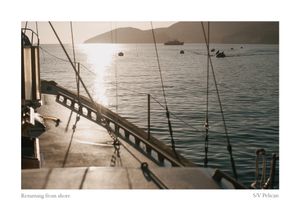 Sunlight glistens on calm bay water as sailboats rest at anchor, viewed from the deck of the sailing vessel SV Pelican.