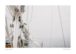 Close-up of a sailboat's white mast with coiled ropes, rigging lines and winches against a calm, misty sea under a pale gray sky.