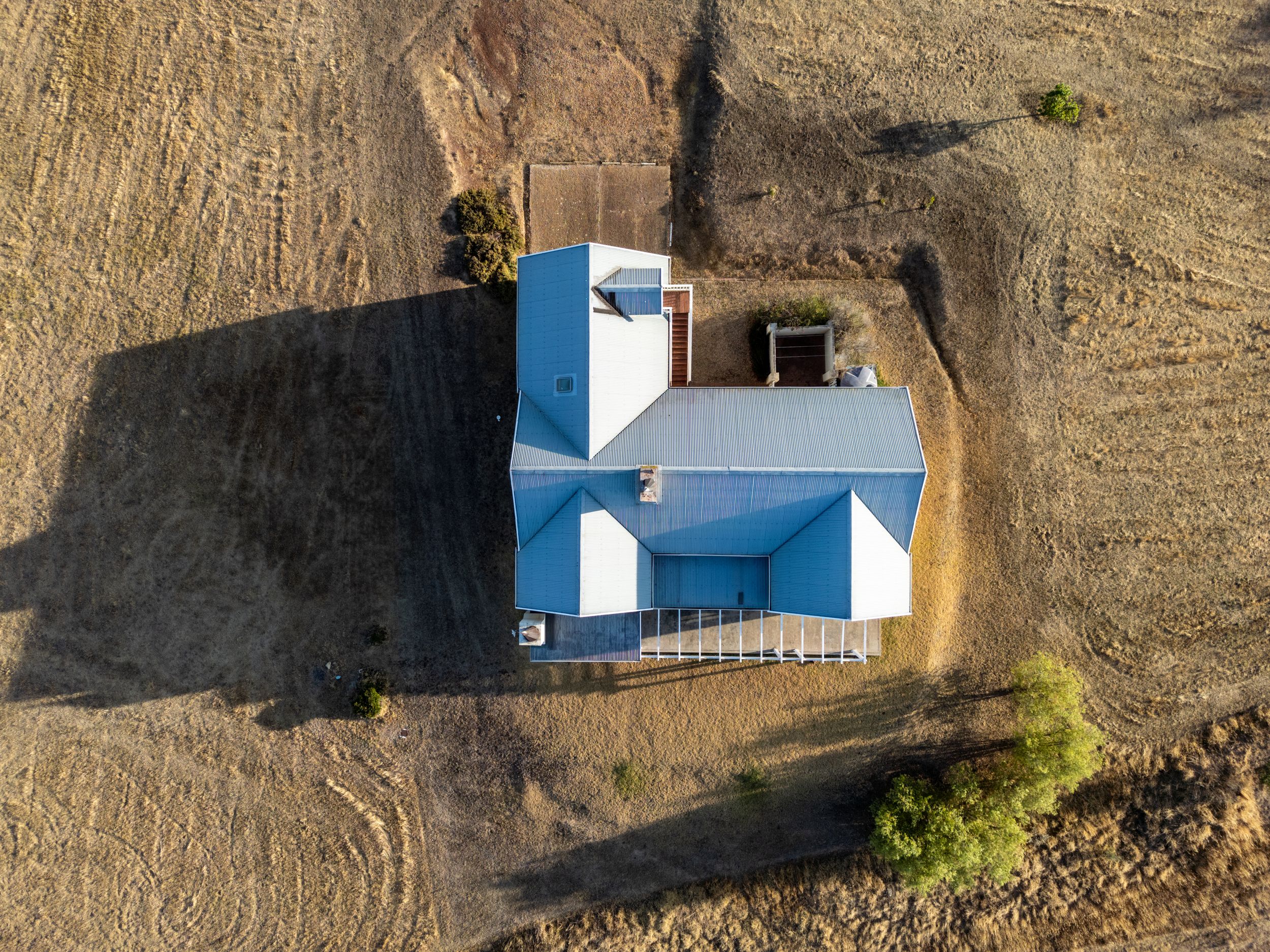 Aerial view of a blue metal roof house on dry rural land with golden grass, a long shadow, and a few trees nearby.