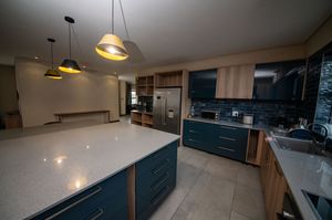 Modern open-plan kitchen with a large gray quartz island, dark teal cabinetry, stainless steel fridge and gold pendant lights.
