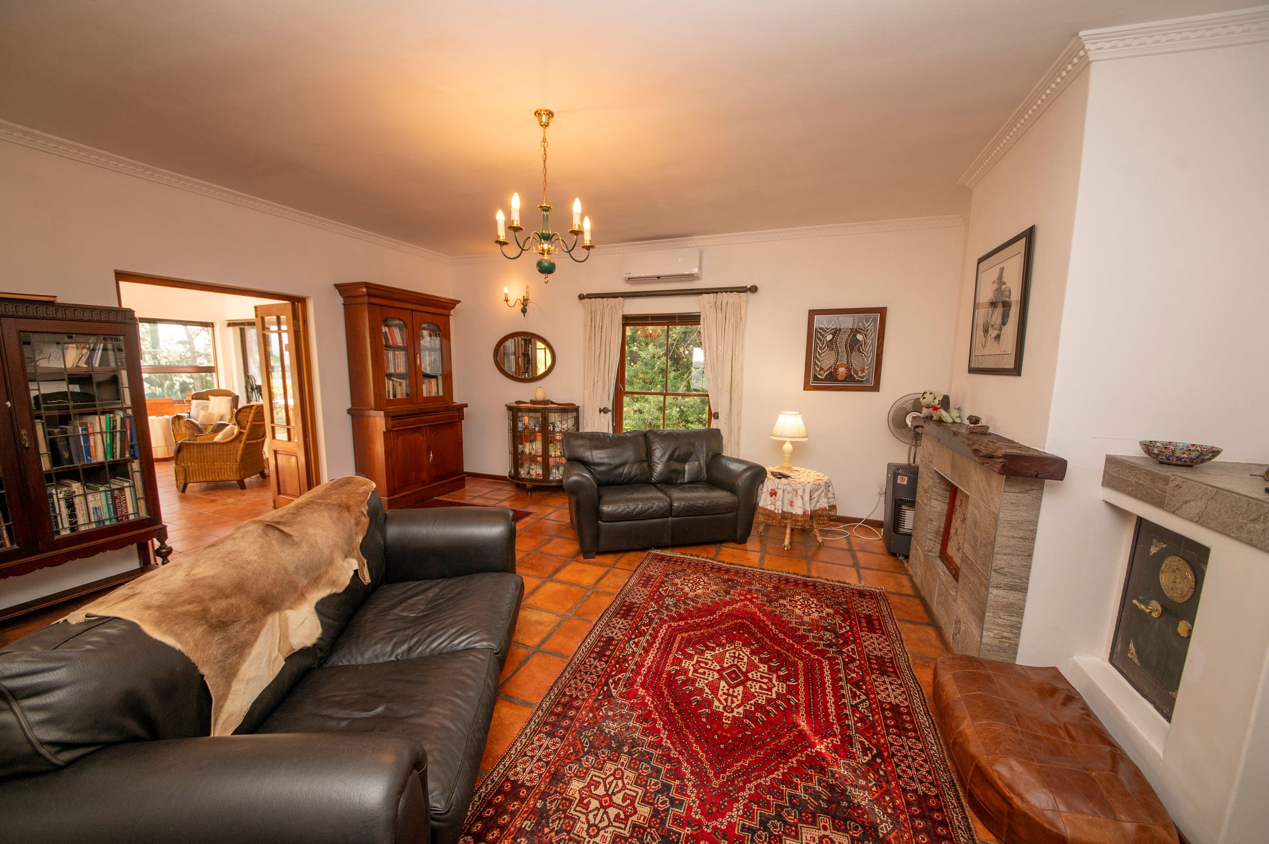 Cozy living room with black leather sofas, red Persian rug, terracotta tile floor, stone fireplace, wooden bookcases and a chandelier.