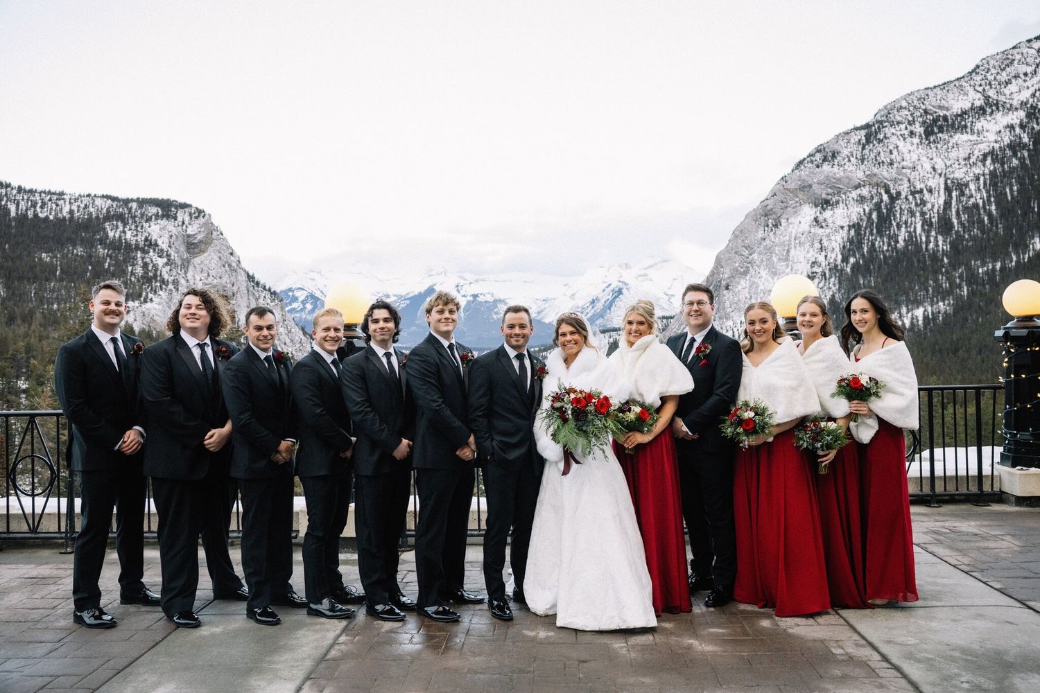 Wedding party in black suits and red dresses with white wraps on a Fairmont Banff Springs terrace with mountain views