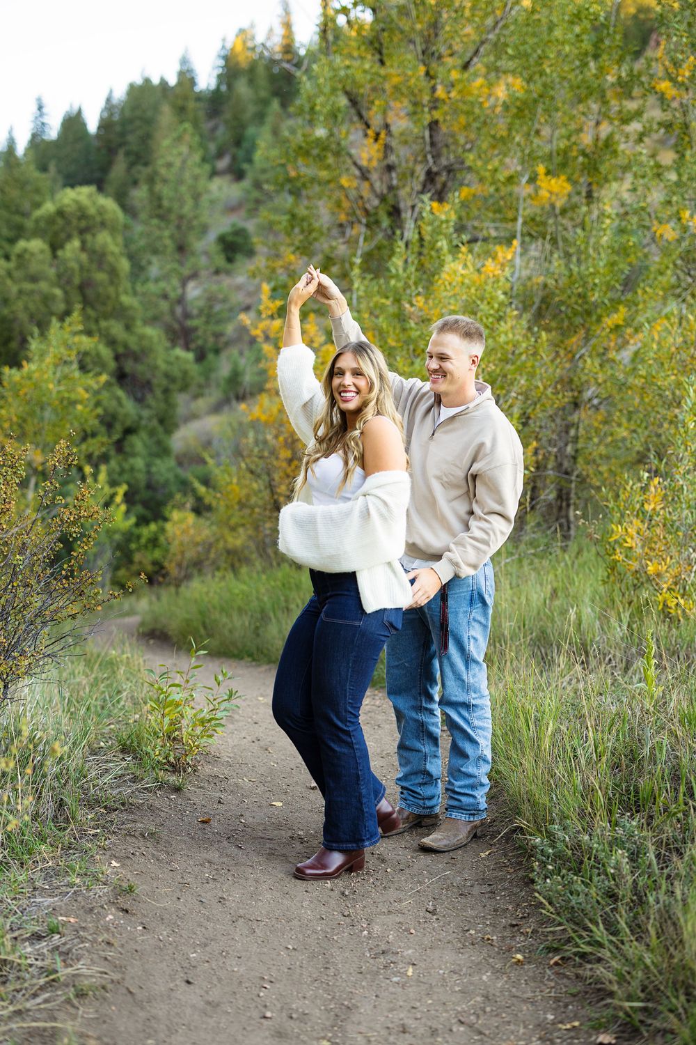 Smiling couple in casual sweaters and jeans dance and twirl on a dirt trail surrounded by green trees and golden autumn foliage.