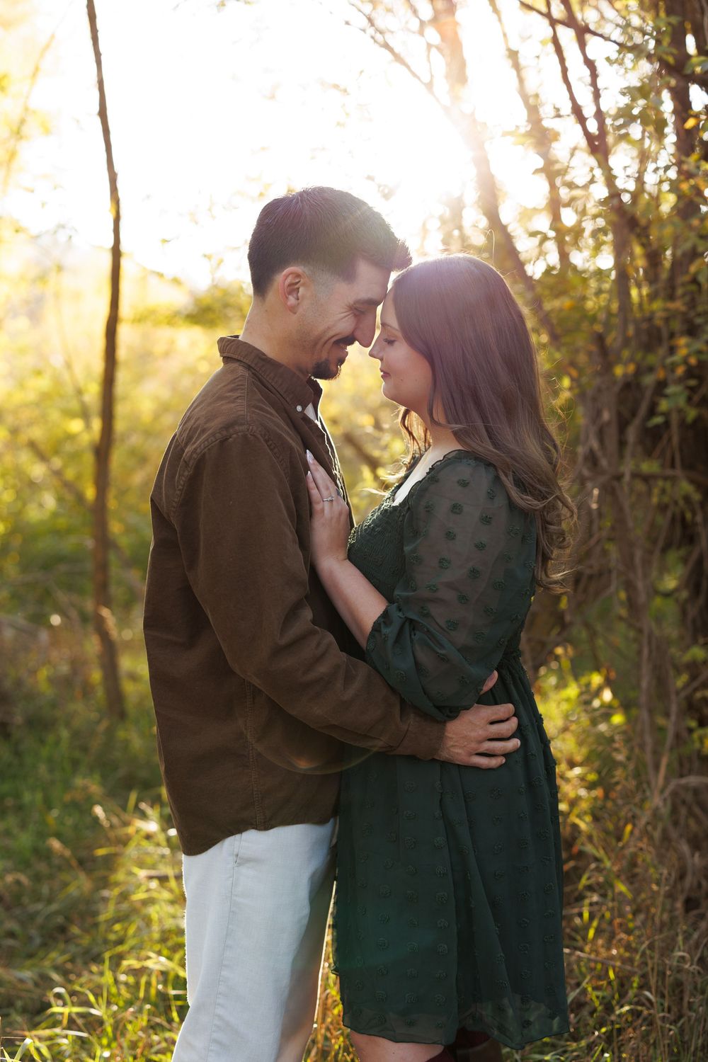 Couple embracing forehead to forehead in golden sunset light, he wears a brown jacket and she a green dress, in a lush wooded field.