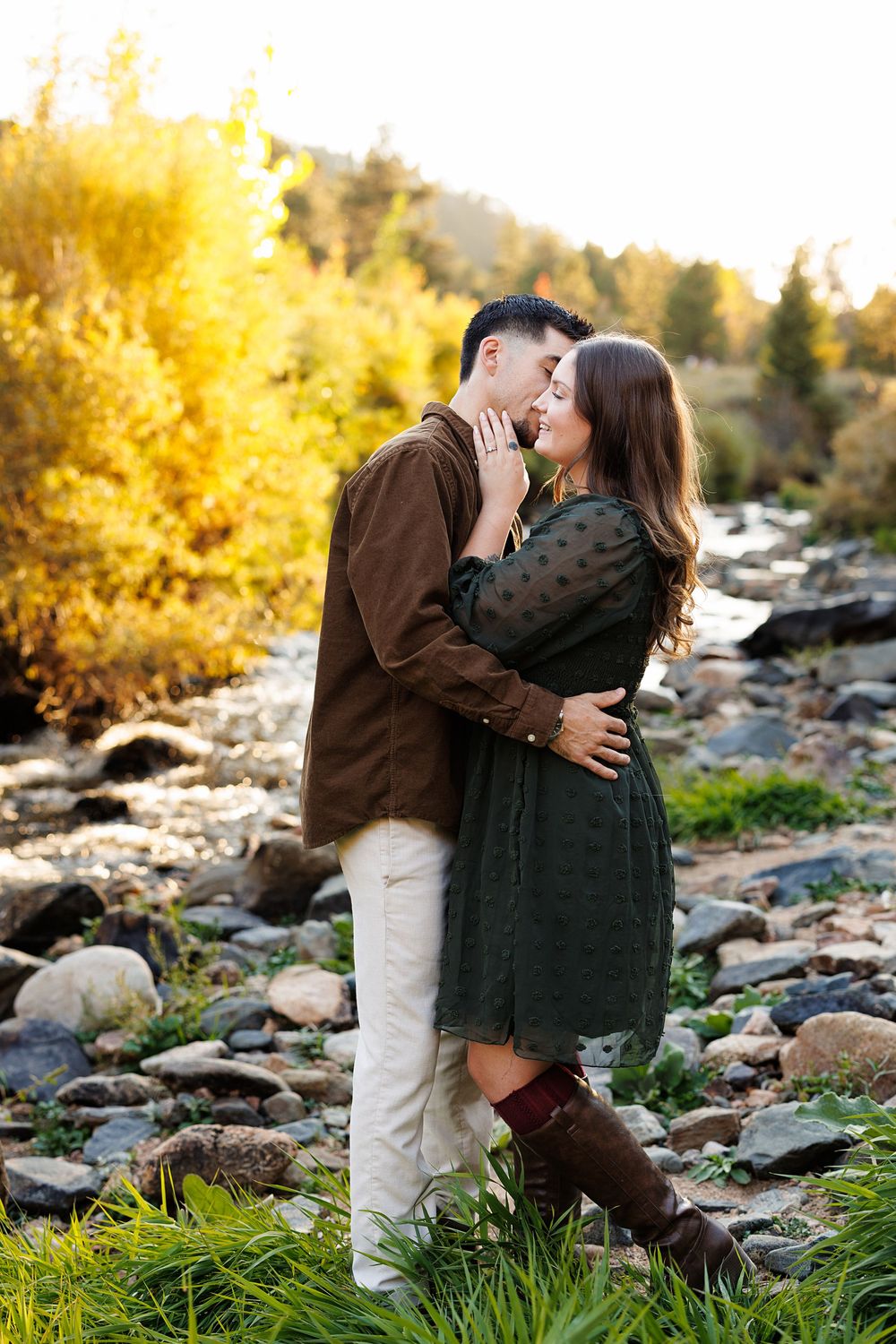 Couple embracing and kissing in tall grass beside a rocky creek, with golden fall trees glowing in warm sunset light.