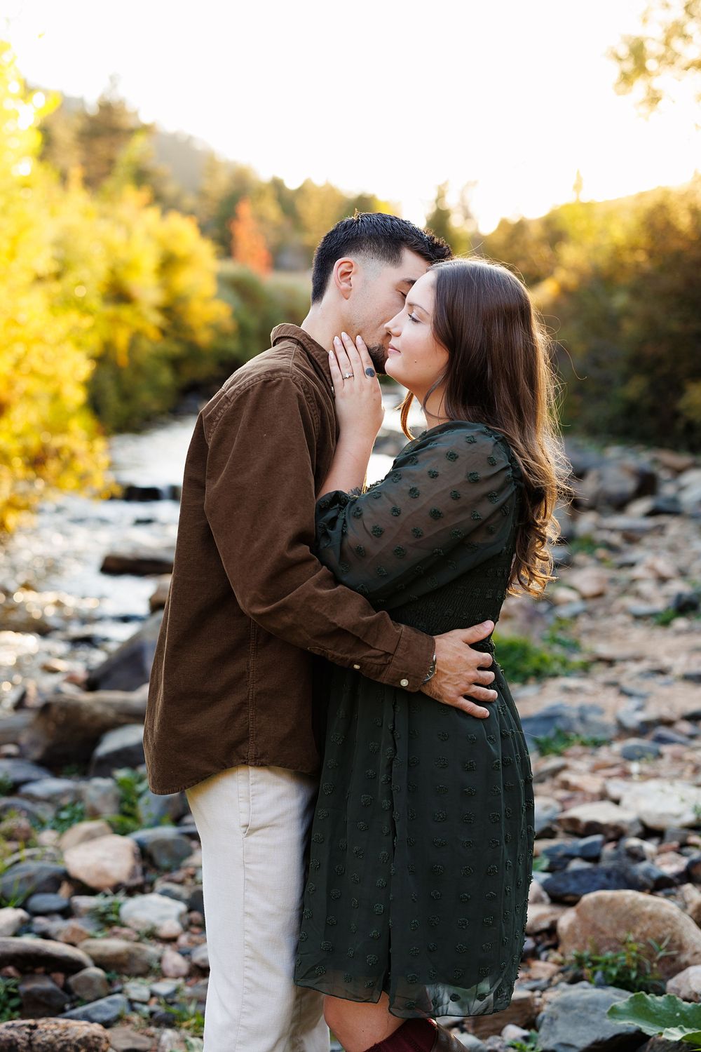 Couple embracing and kissing on a rocky riverbank surrounded by golden autumn foliage during an outdoor engagement photo session.