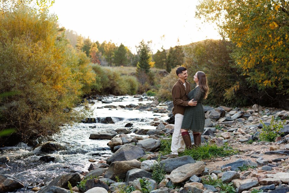 Couple embracing on a rocky riverbank surrounded by golden autumn foliage during an engagement photo session.