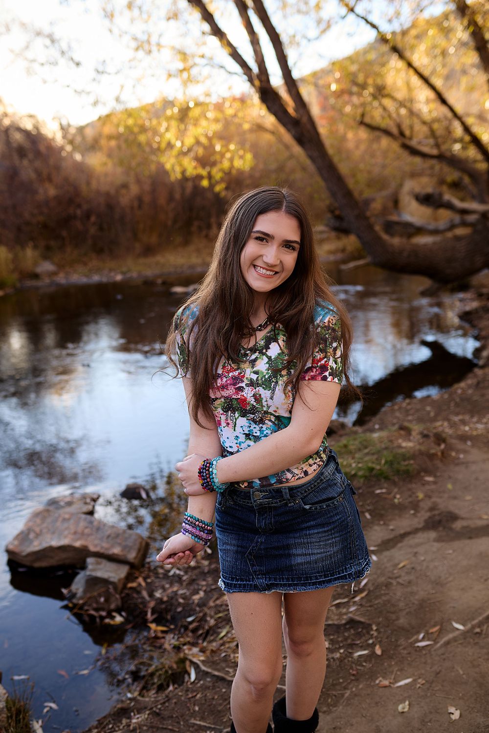 Smiling young woman with long brown hair in a floral top and denim skirt stands on a dirt path beside a calm creek in autumn.