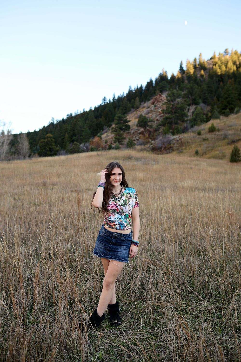 Young woman in a floral top, denim skirt and black boots poses in a golden grassy meadow with forested hills behind her.