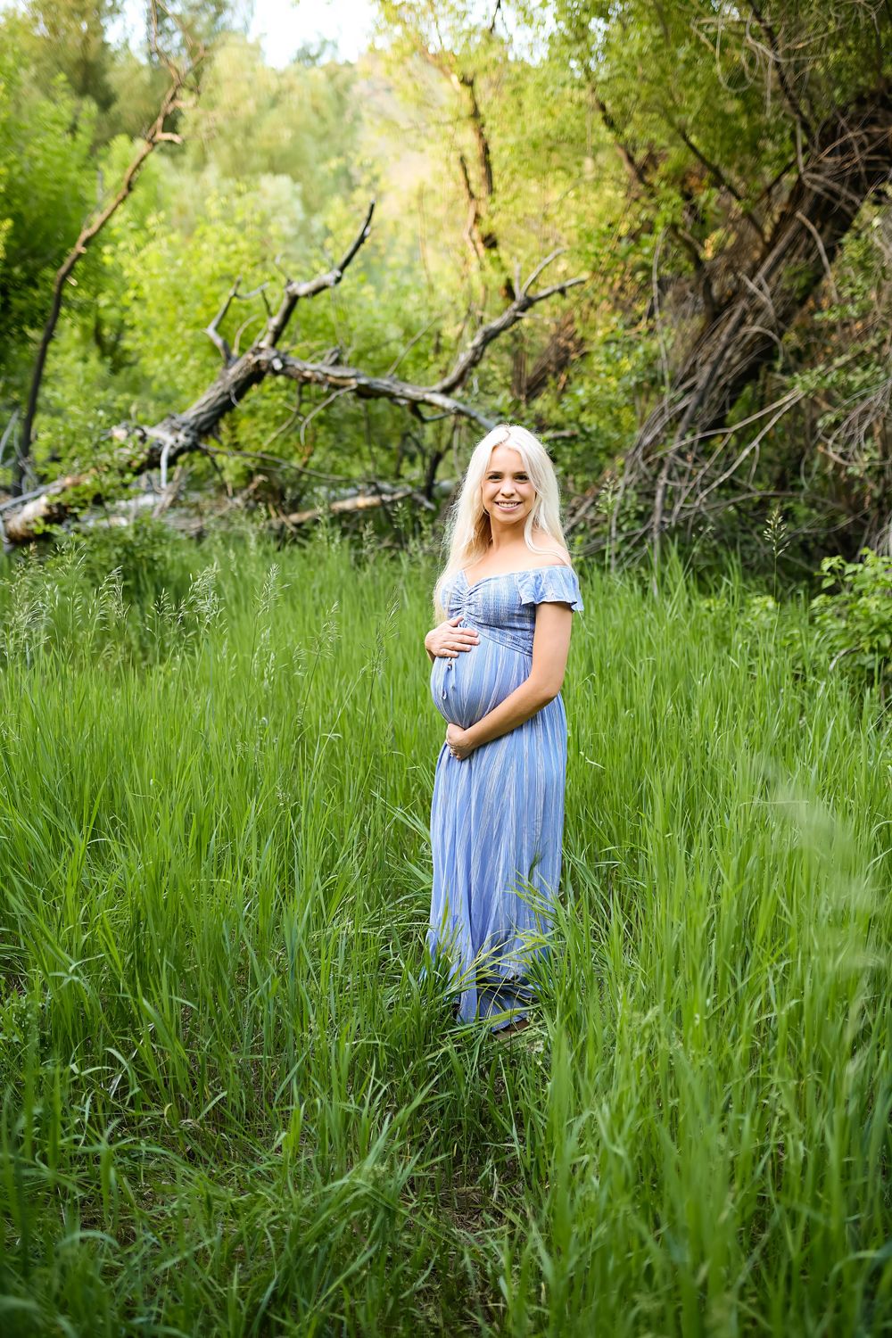 Pregnant woman with long blonde hair in a blue off-the-shoulder dress cradles her baby bump while standing in tall green grass near trees.