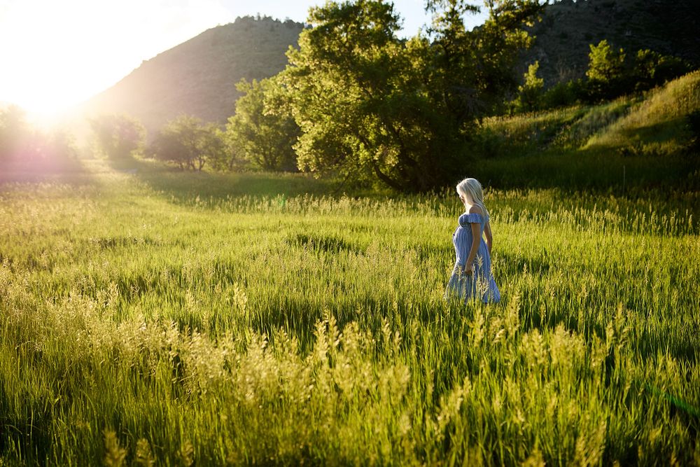 Woman in a light blue dress walks through a tall green grass meadow at sunset, with trees and rolling hills glowing in warm golden light.