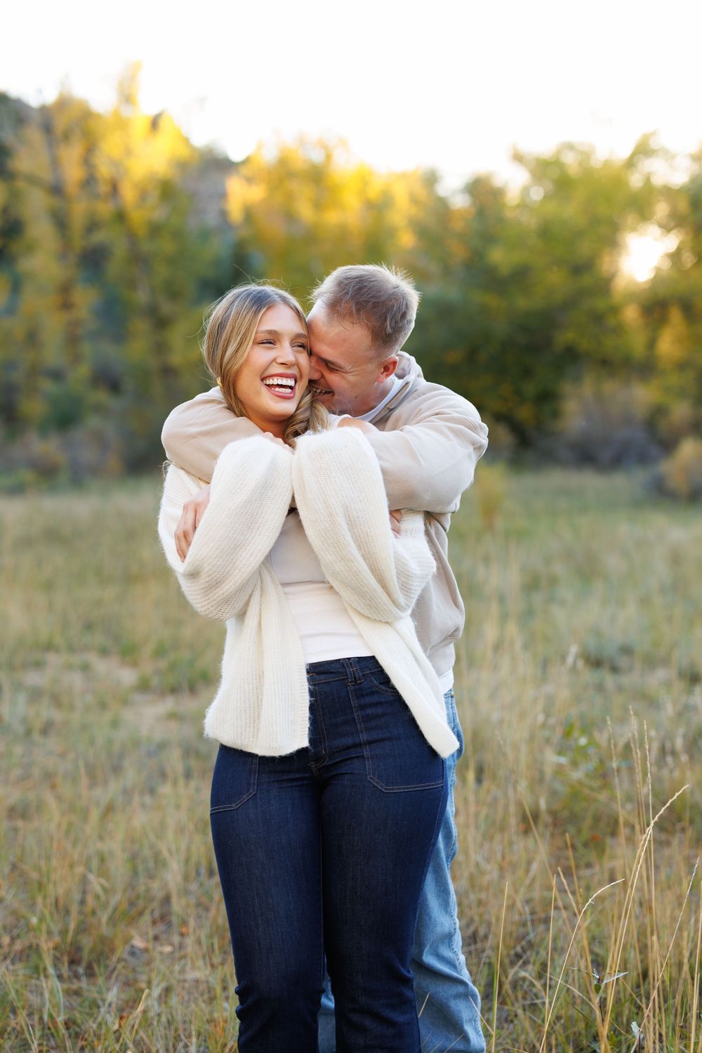 Smiling woman in a cream cardigan and jeans is hugged from behind by a man in a grassy field with golden autumn trees.