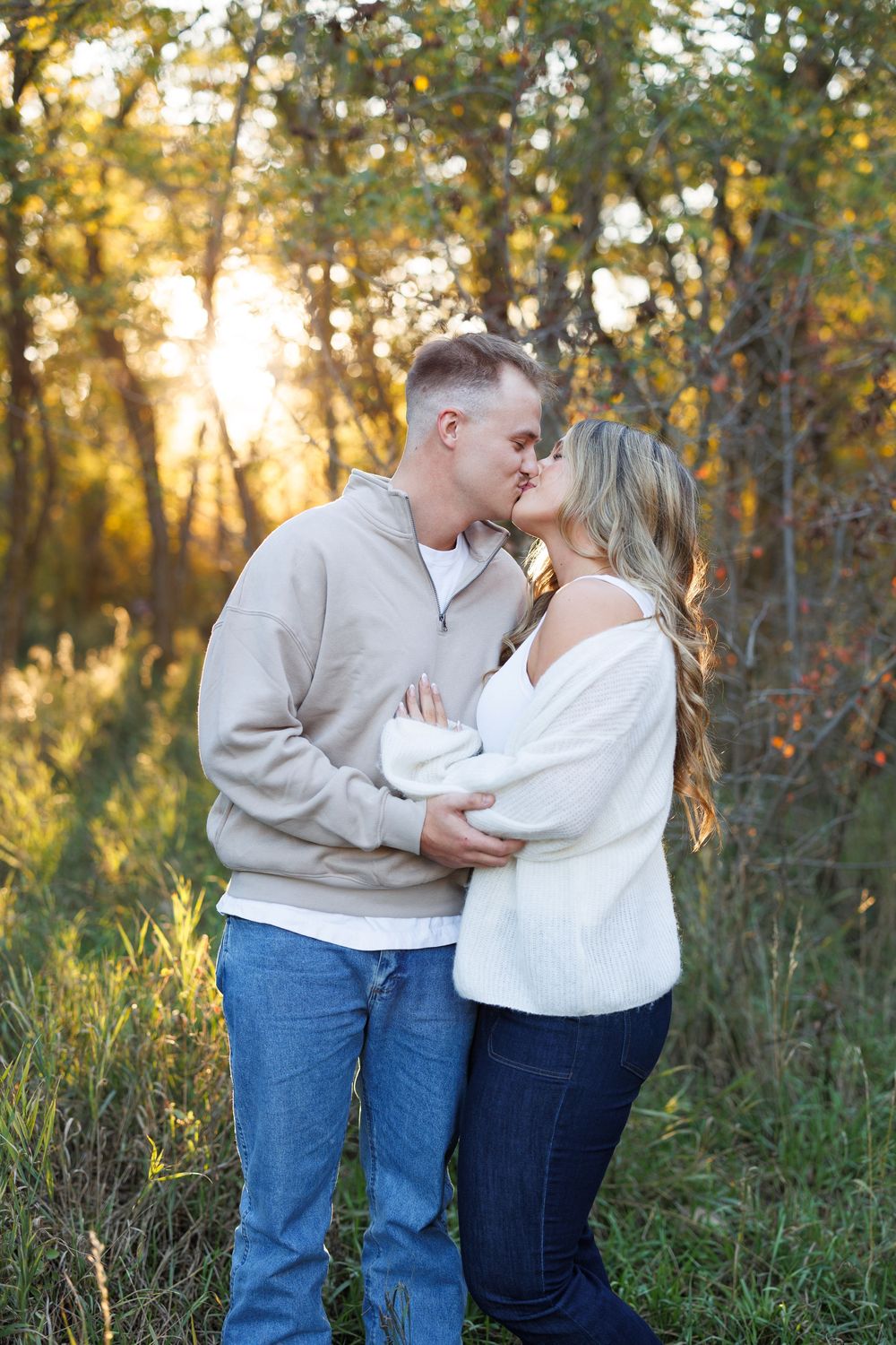 Couple in cozy sweaters and jeans share a kiss in a golden-lit autumn forest with warm sunlight filtering through the trees.