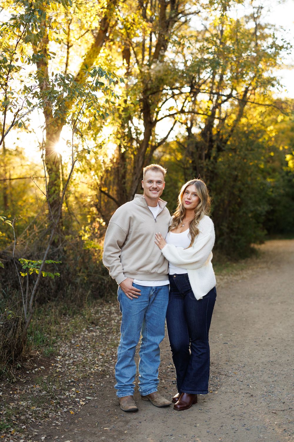 Smiling couple standing close together on a dirt path in a sunlit autumn forest, wearing casual sweaters, jeans and boots.