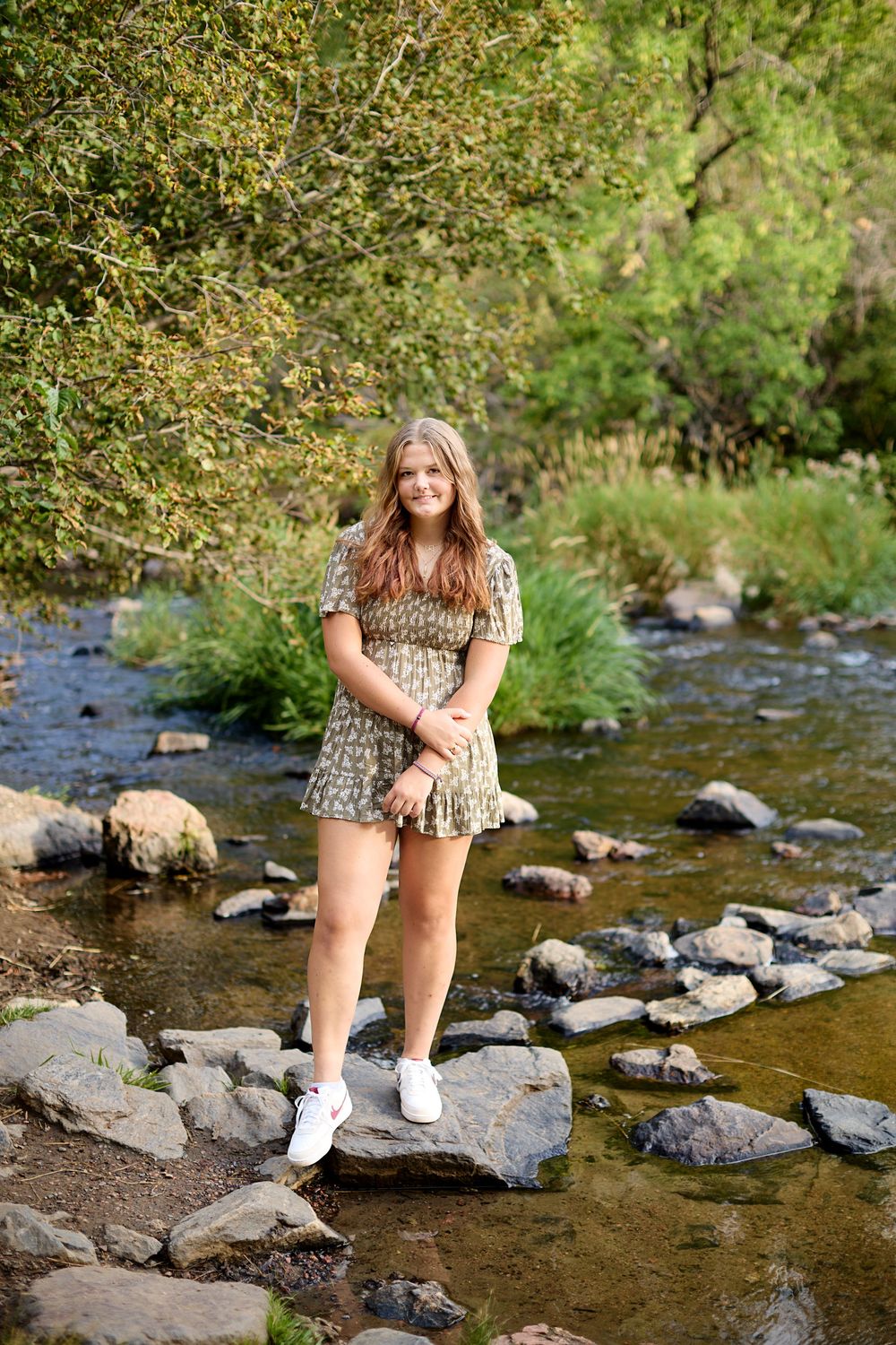 Young woman in a floral dress and white sneakers stands on a large rock in a shallow, sunlit rocky stream surrounded by green trees.