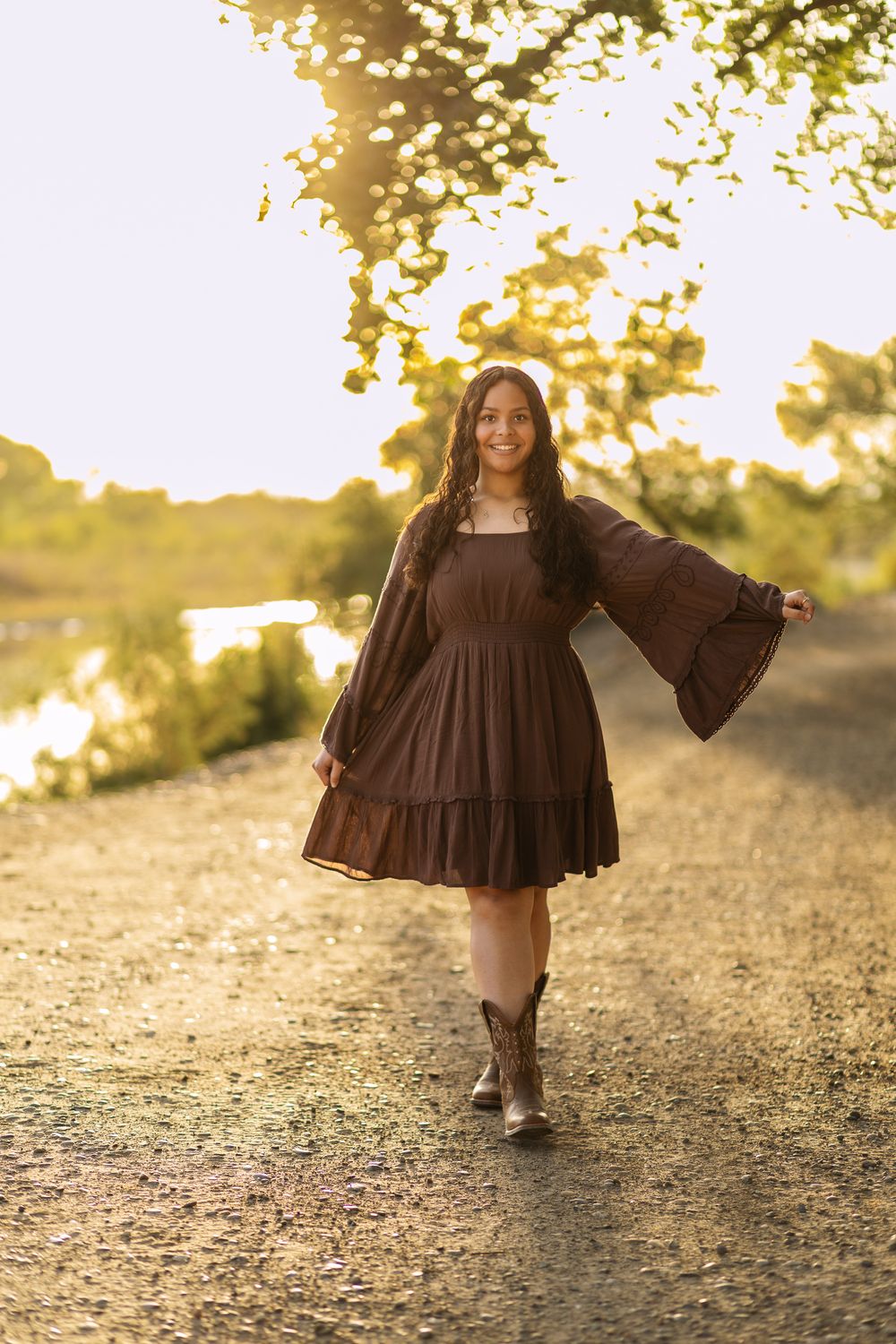 Smiling young woman in a brown flowy dress and cowboy boots walks along a gravel path in warm golden sunset light.