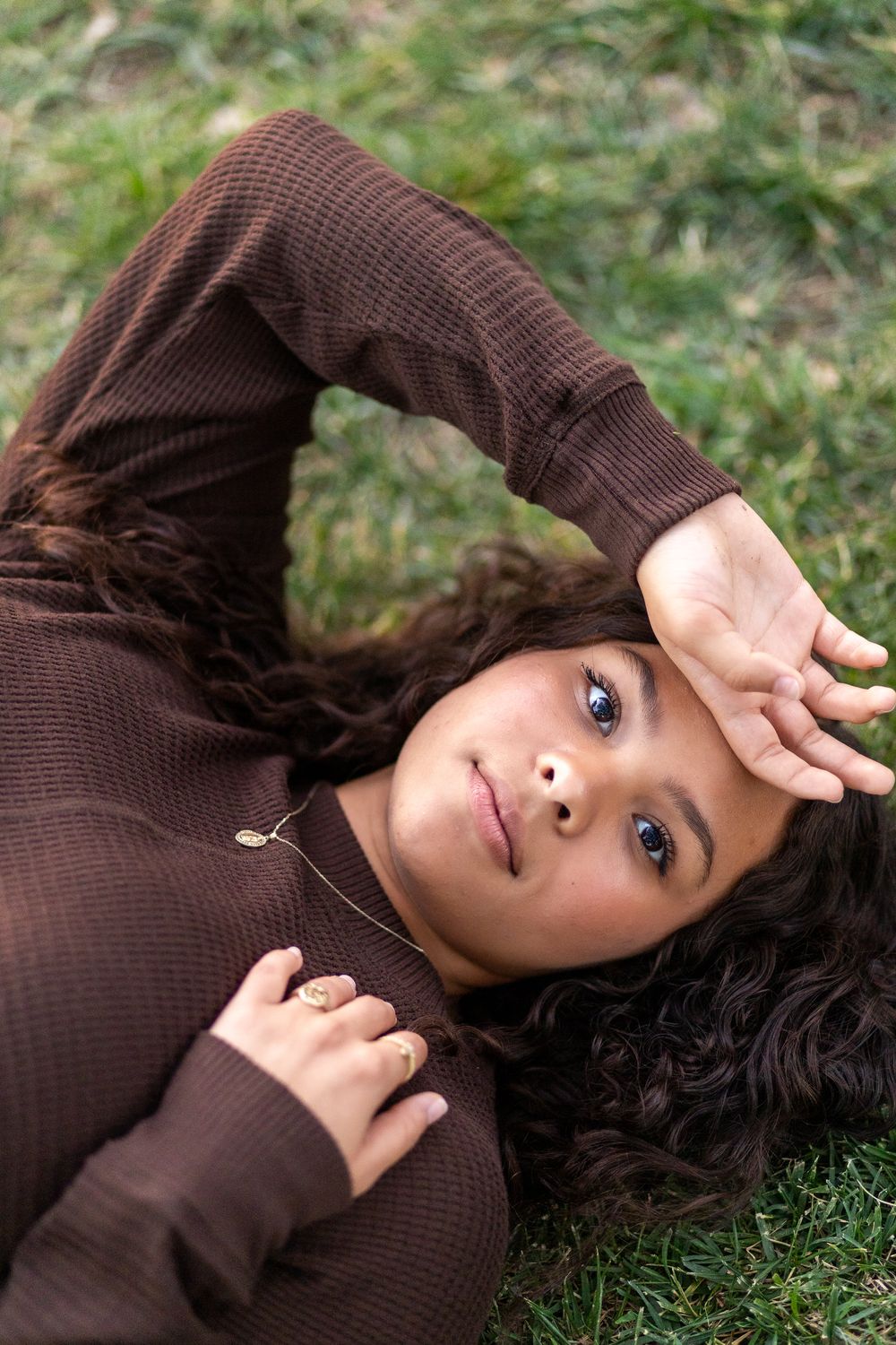 Young woman with dark curly hair rests on green grass, one arm raised over her forehead in a calm portrait.