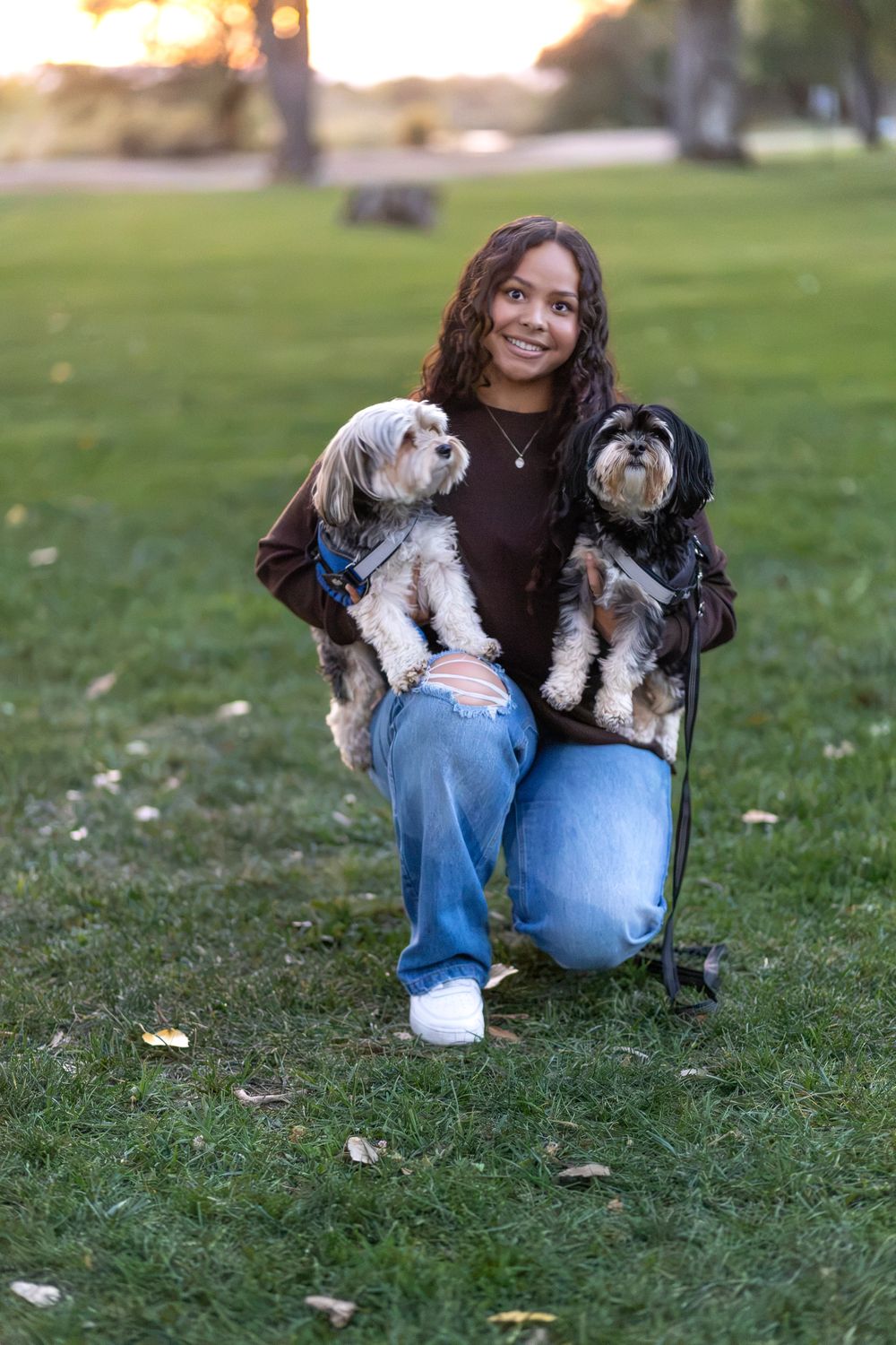 Happy young woman kneeling on grass in a park, holding two small fluffy dogs in her arms during a golden hour portrait.