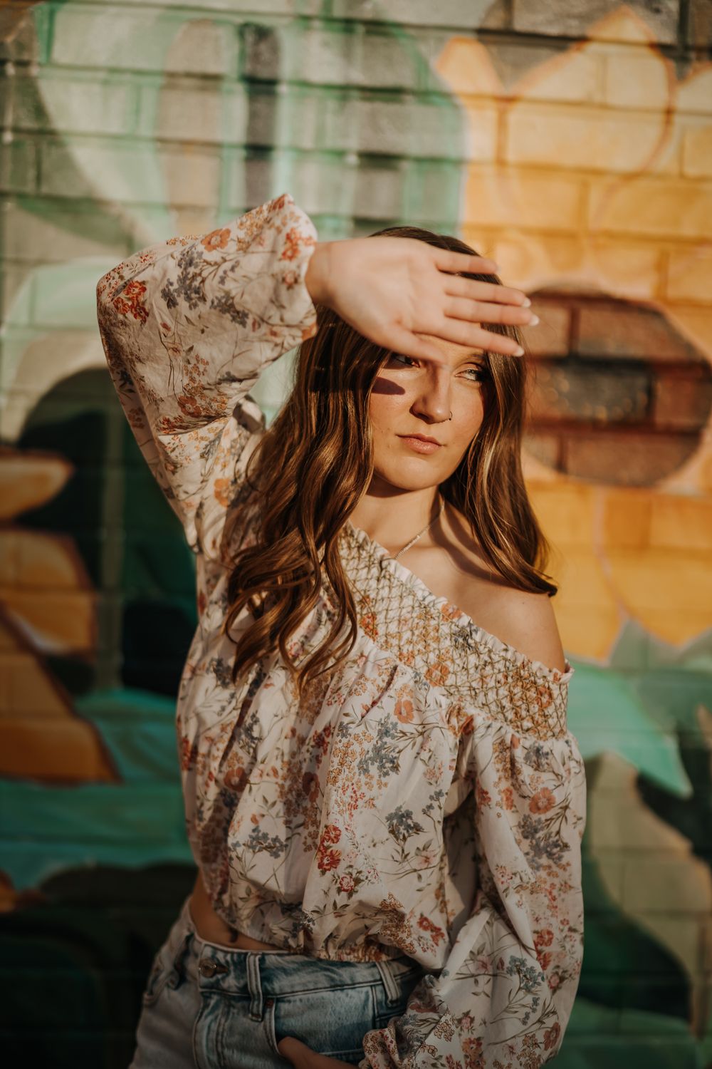 Woman in a floral off-shoulder top shades her eyes with her hand in front of a colorful brick wall mural.