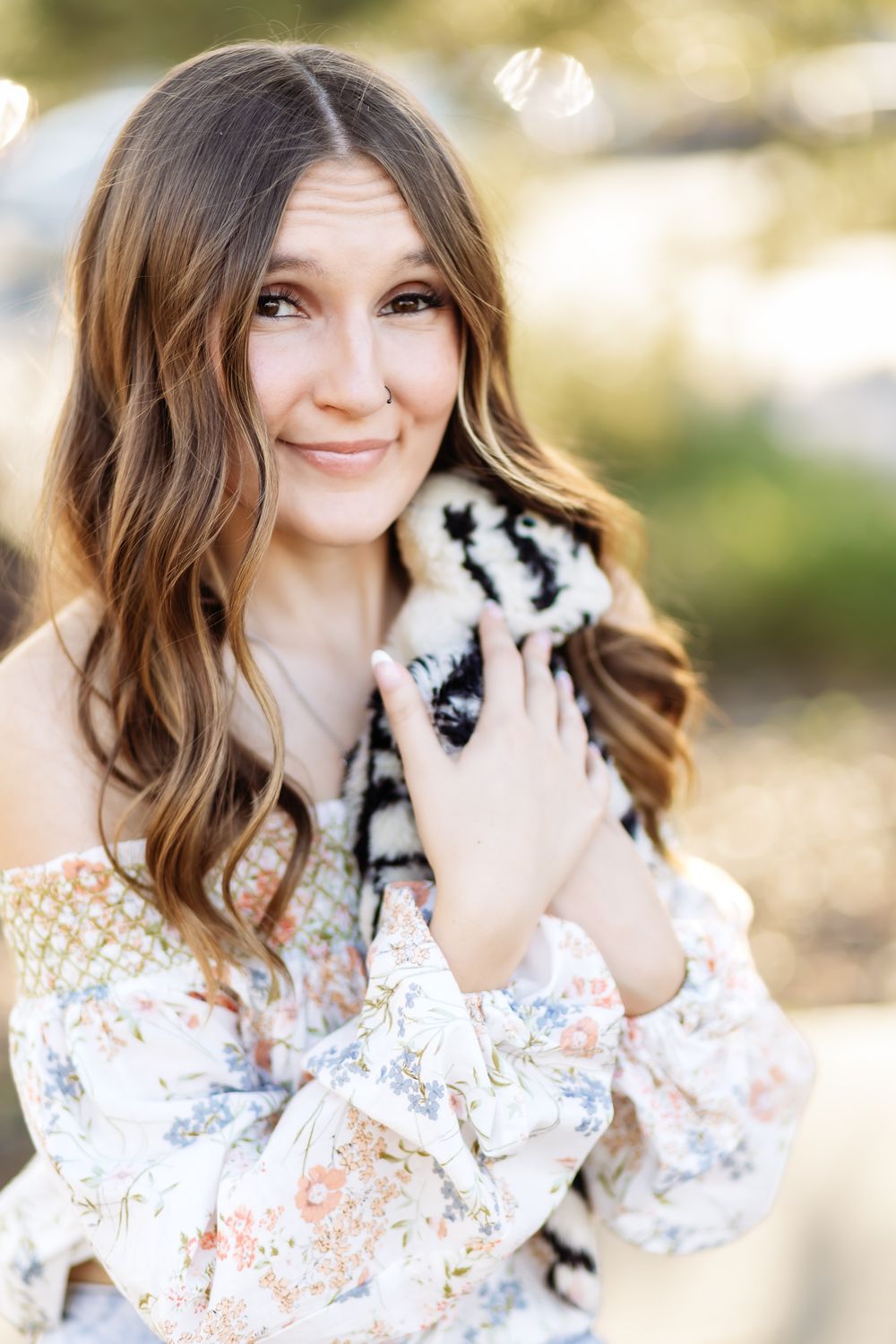 Smiling young woman with long wavy brown hair in a floral off-shoulder top cuddling a small black and white fluffy animal outdoors.