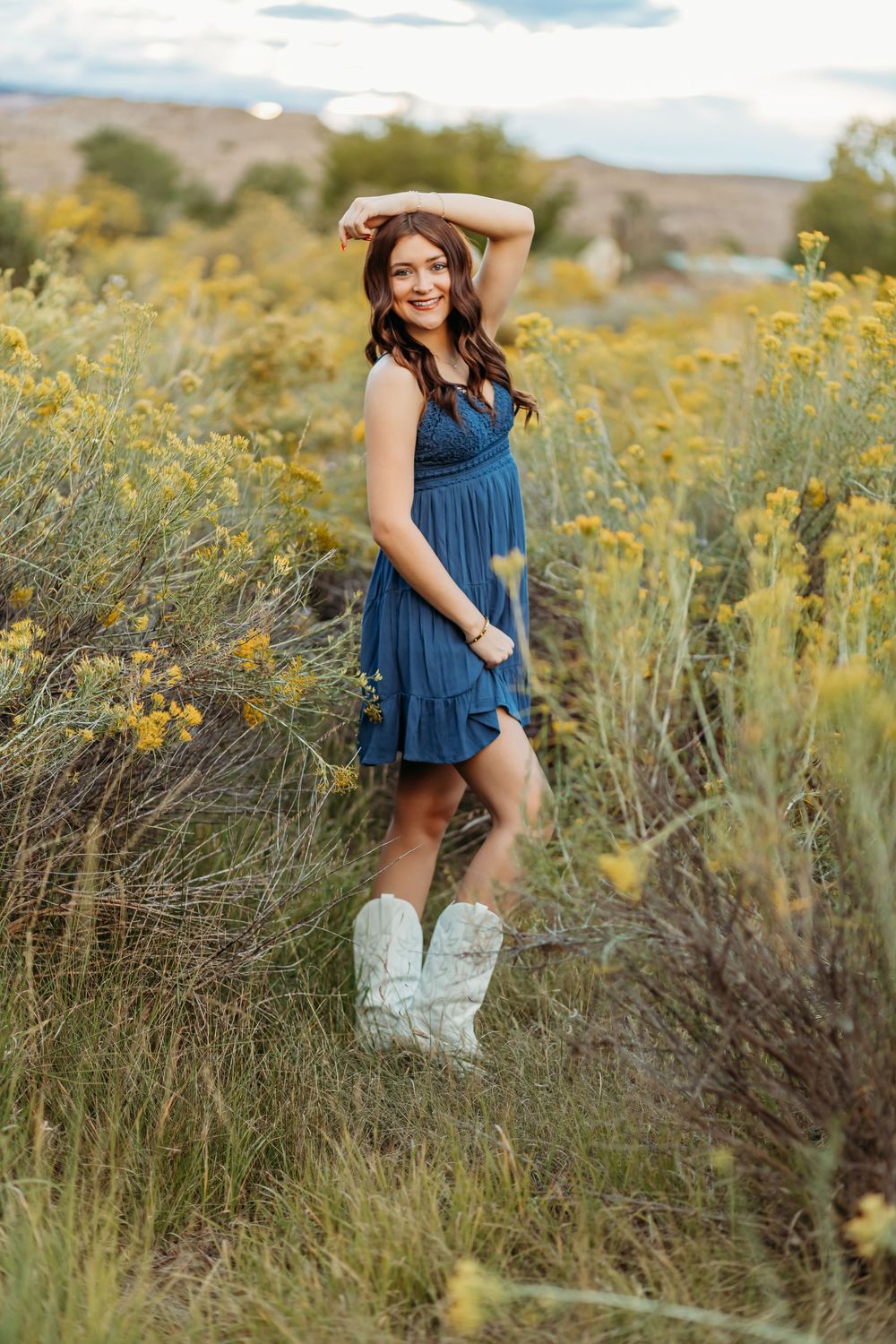 Woman in a blue dress and white cowboy boots poses with one hand on her head in a field of yellow wildflowers.