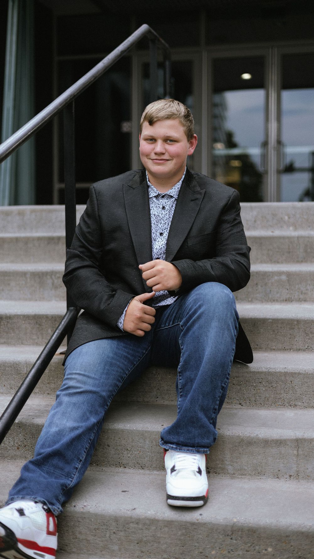 Smiling young man in a blazer, jeans and red-and-white sneakers sitting on concrete steps beside a black metal railing.