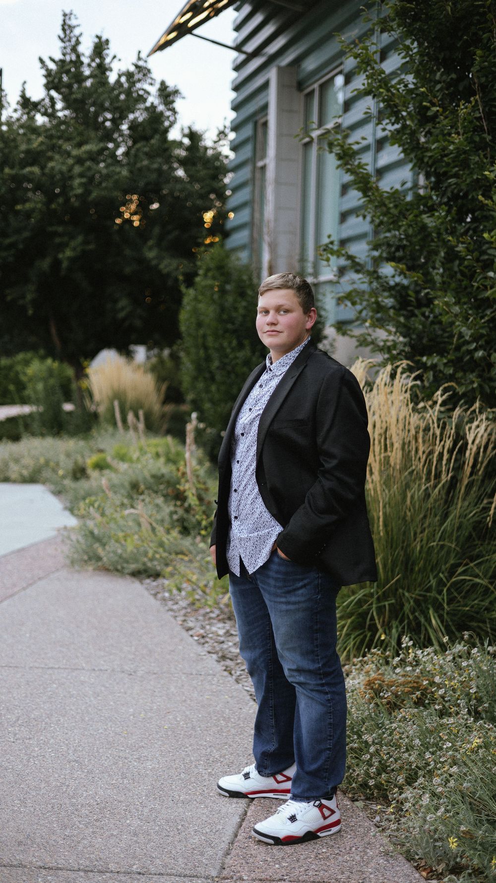 Young man in a black blazer, patterned shirt and jeans standing on a garden sidewalk lined with ornamental grasses.