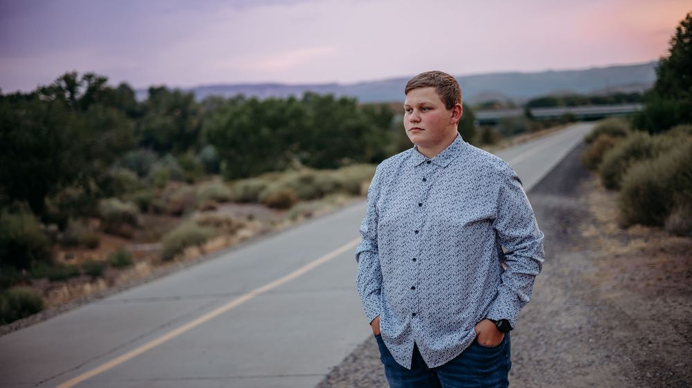 Young man in a blue checkered shirt standing along a quiet paved trail with a pink and purple sunset sky behind him.