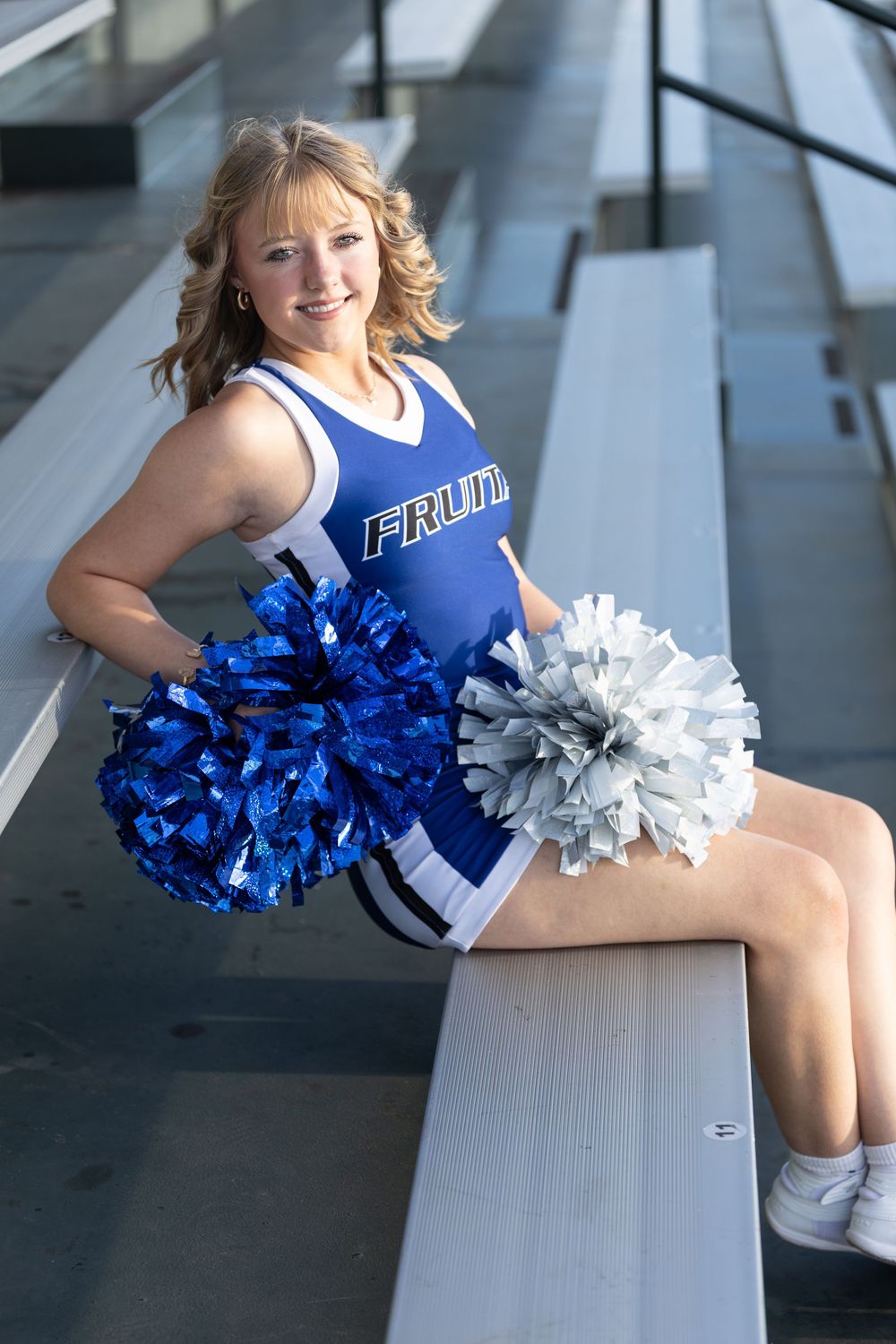 Cheerleader in a blue uniform sits on stadium bleachers holding blue and silver pom-poms and smiling at the camera.