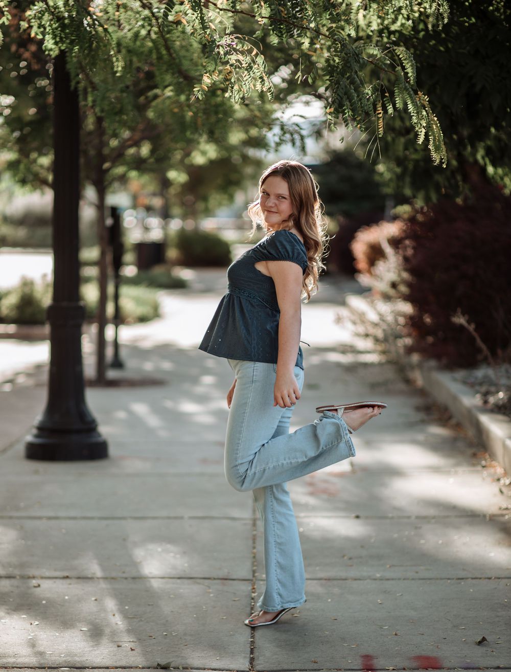 Young woman in a blue top and flared jeans poses with one foot raised on a tree-lined sidewalk.