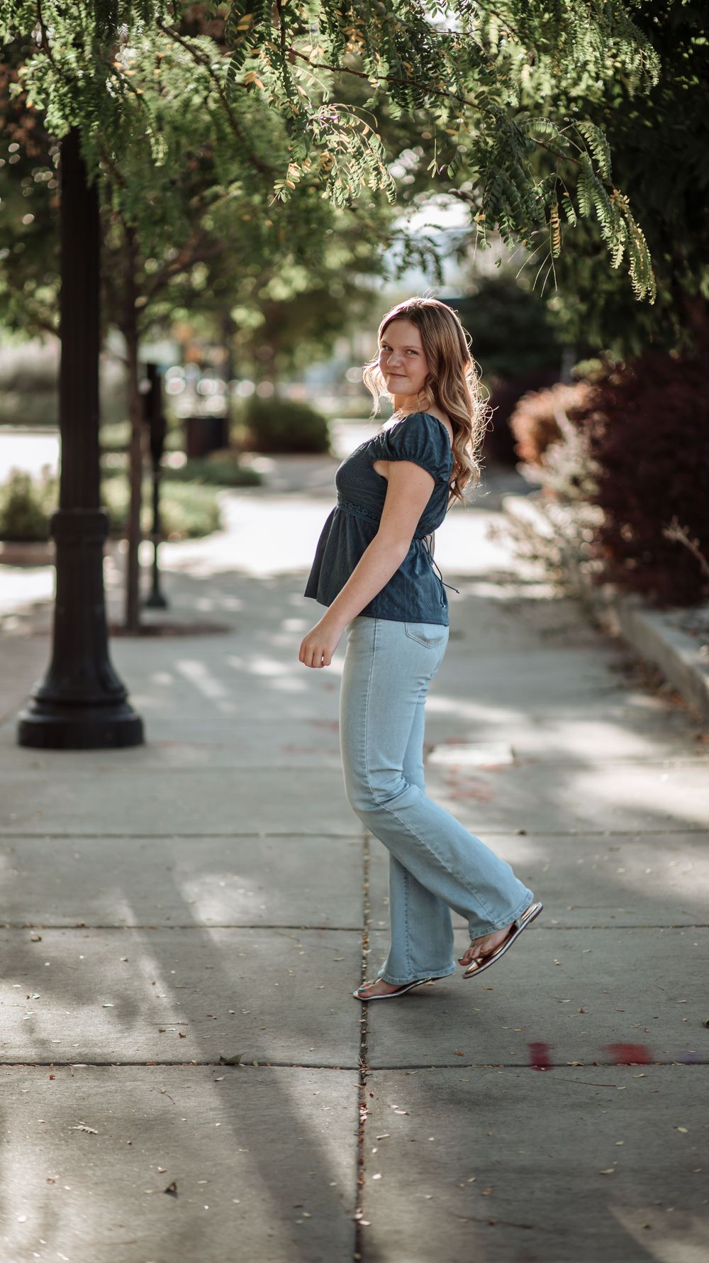 Young woman in a blue blouse and light jeans walks along a sunlit tree-shaded sidewalk while looking back.