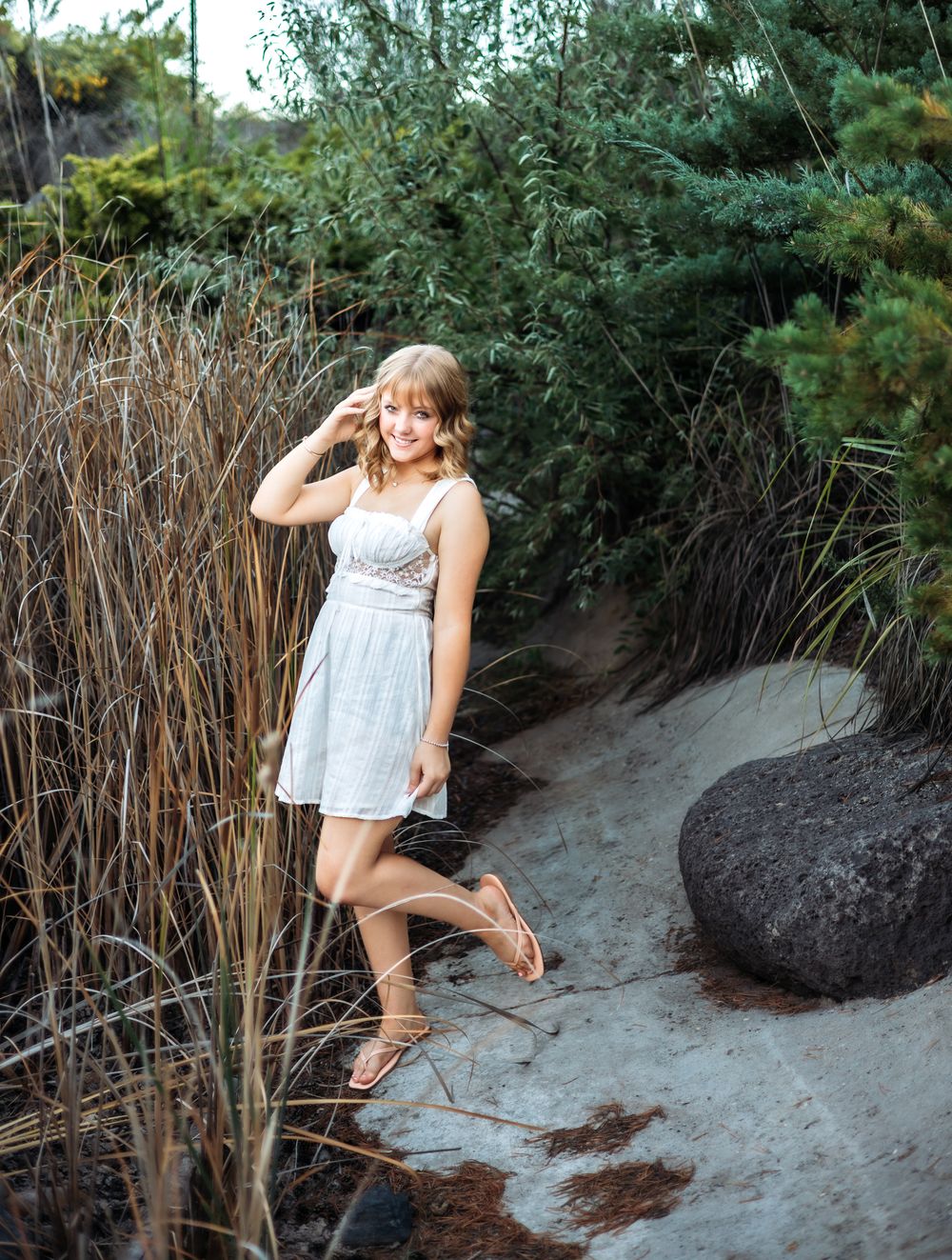 Smiling young woman in a white sundress poses on a sandy path beside tall grasses, evergreen trees and a dark boulder.