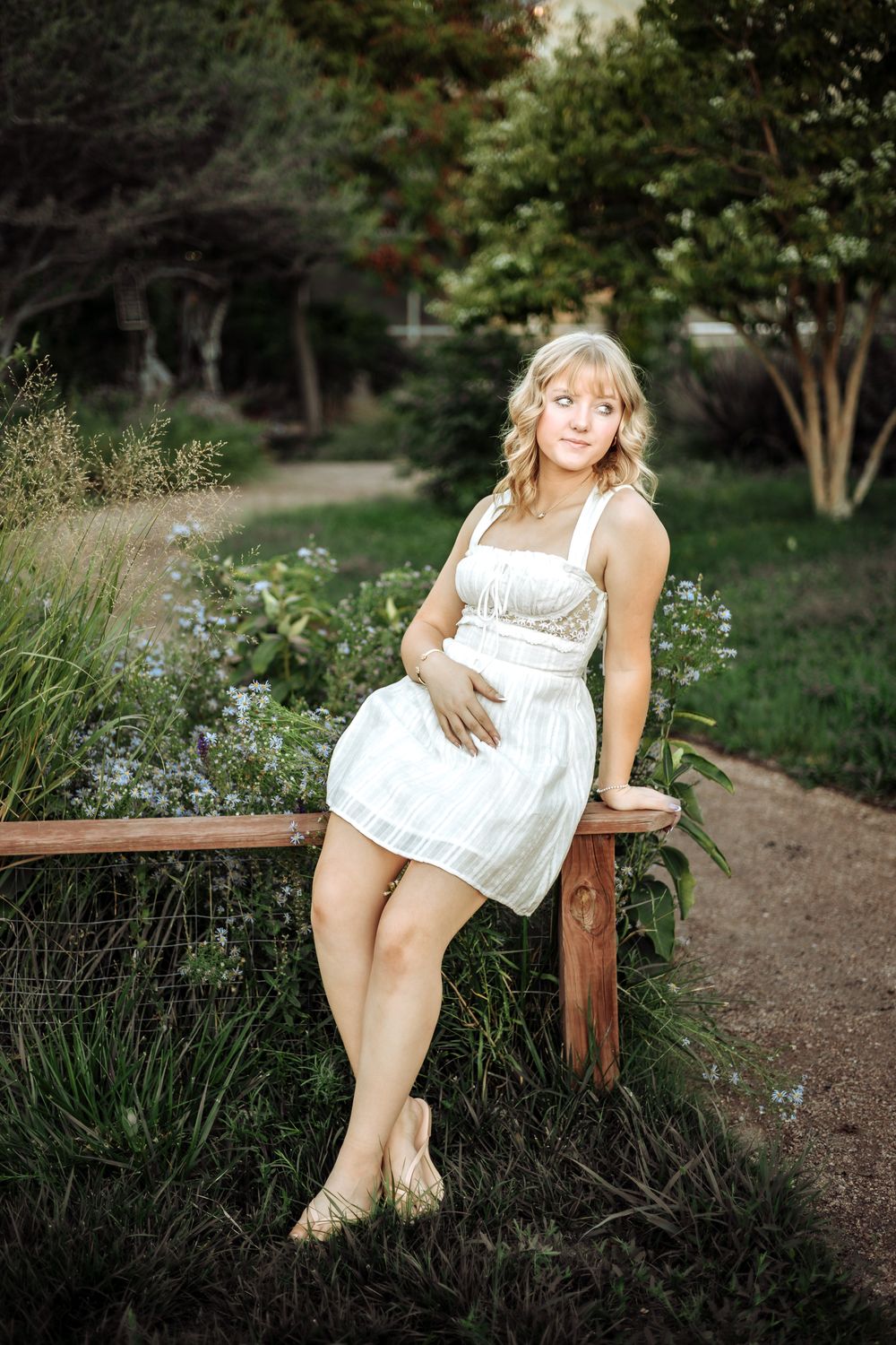 Blonde young woman in a white sundress sits on a wooden fence rail in a lush garden with flowers and a gravel path.