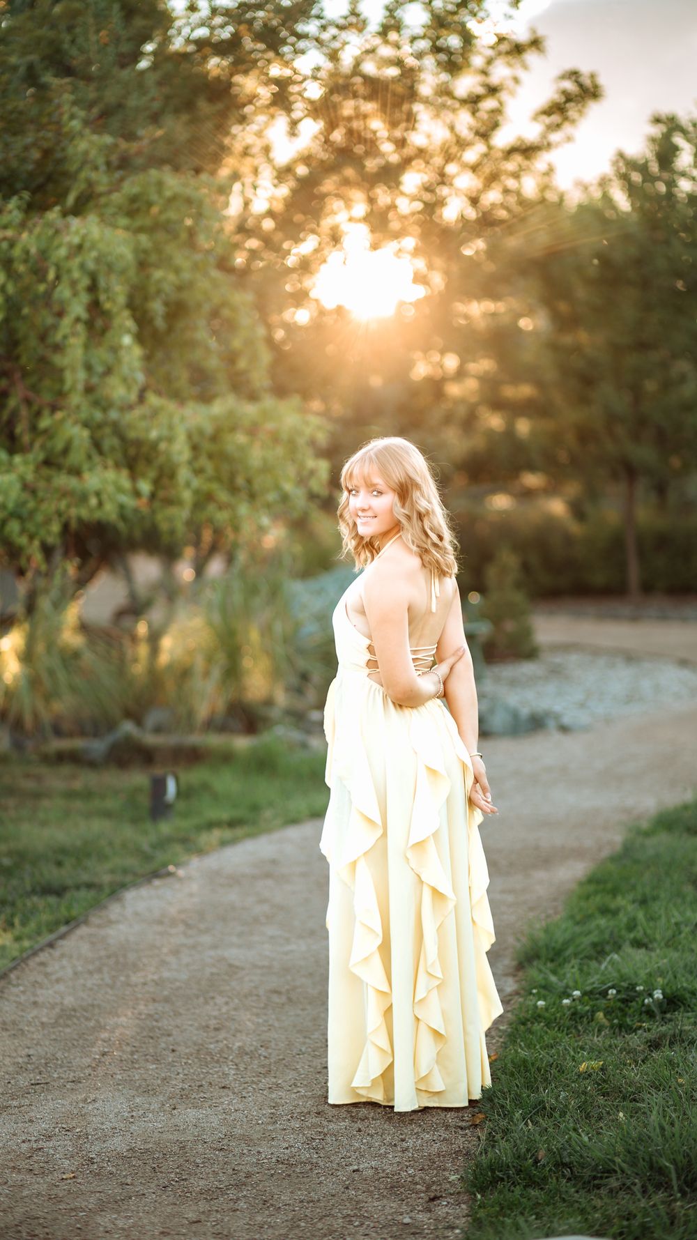 Woman in a flowing pale yellow ruffled gown glances over her shoulder on a gravel path as the golden sun sets through the trees.