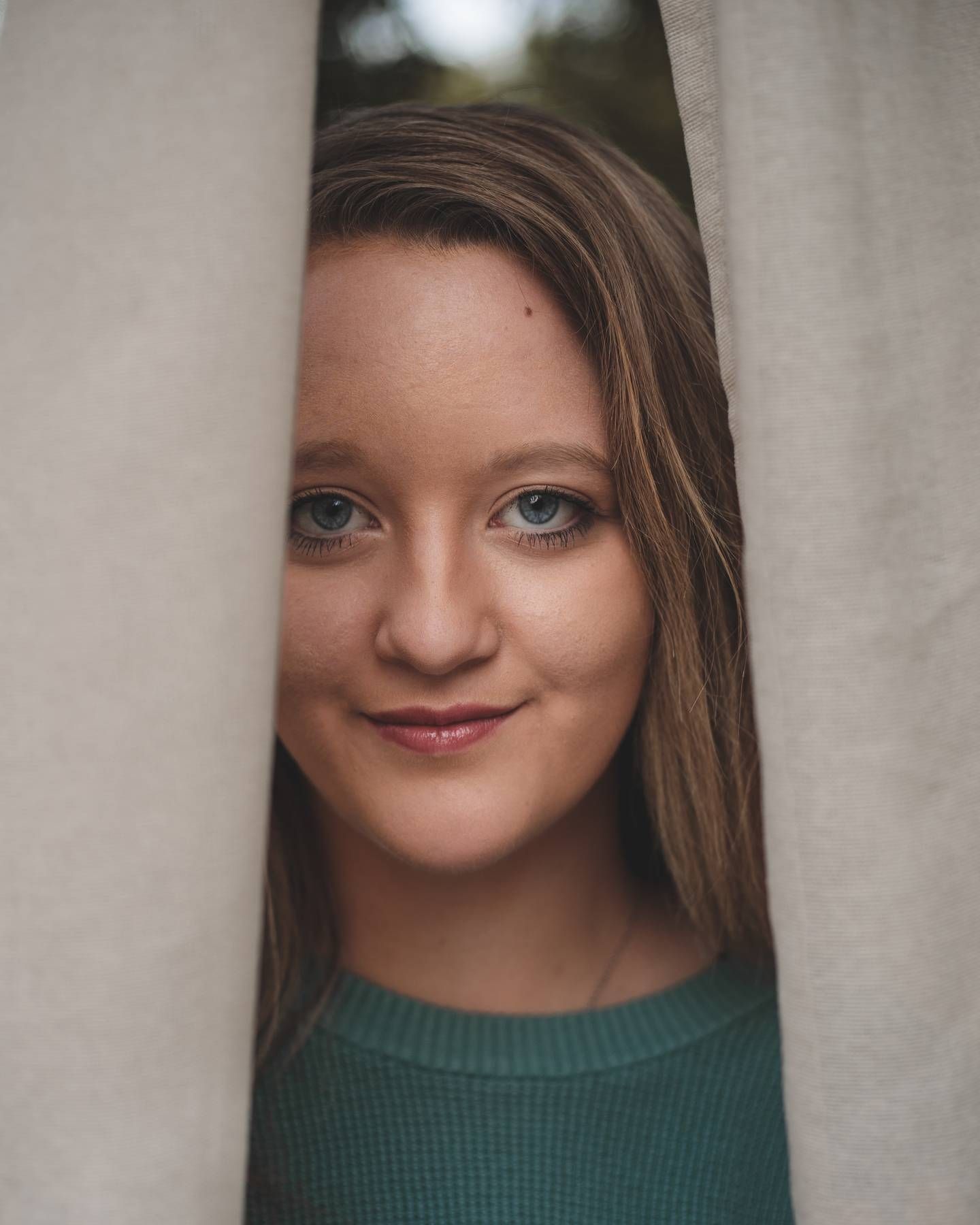 A young woman with blue eyes and a teal sweater peeks out between two beige curtains with a gentle smile.