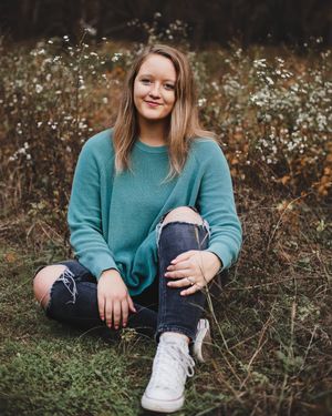 A young woman in a teal sweater and ripped jeans sits smiling on the grass in a field of wildflowers.